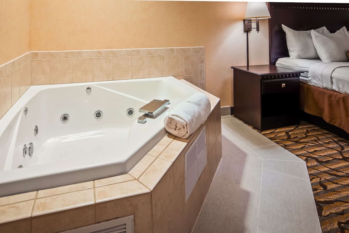 Un baño de hotel con una bañera grande, una mesita de noche y una cama con almohadas.
