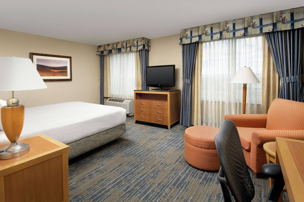 A hotel room with a bed, a desk with a lamp, a television, an ottoman, and a chair.