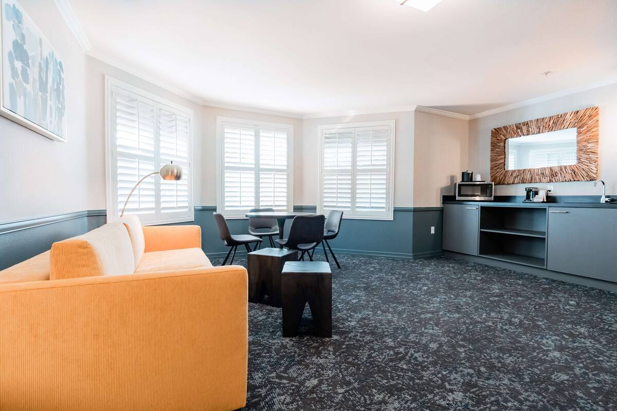 Una habitación de hotel moderna con sofá, una mesa de comedor con sillas, kitchenette y ventanas amplias con persianas.