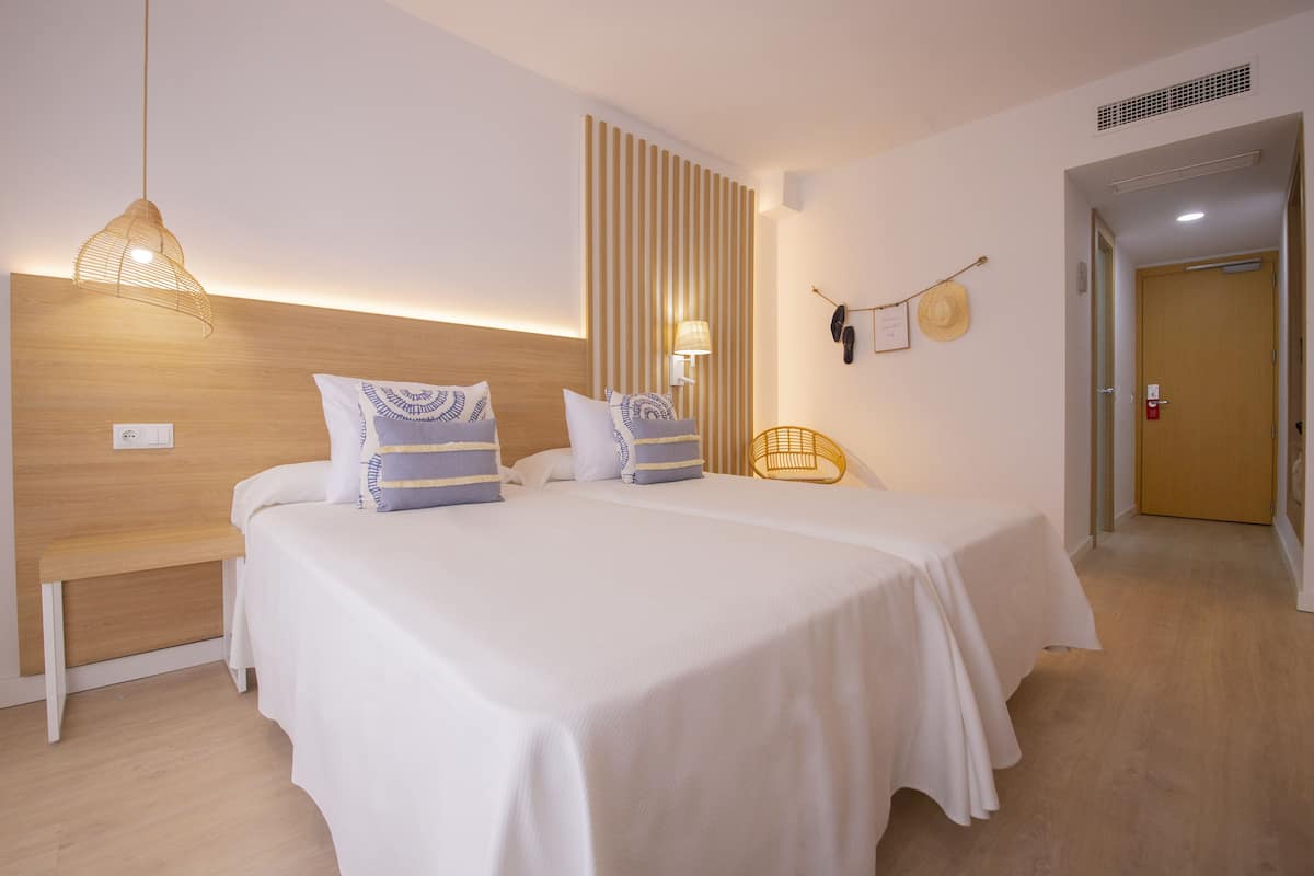 Habitación de hotel con dos camas, cabecero de madera, mesita de noche, lámpara colgante, lámpara de pared y una puerta con cartel de seguridad.