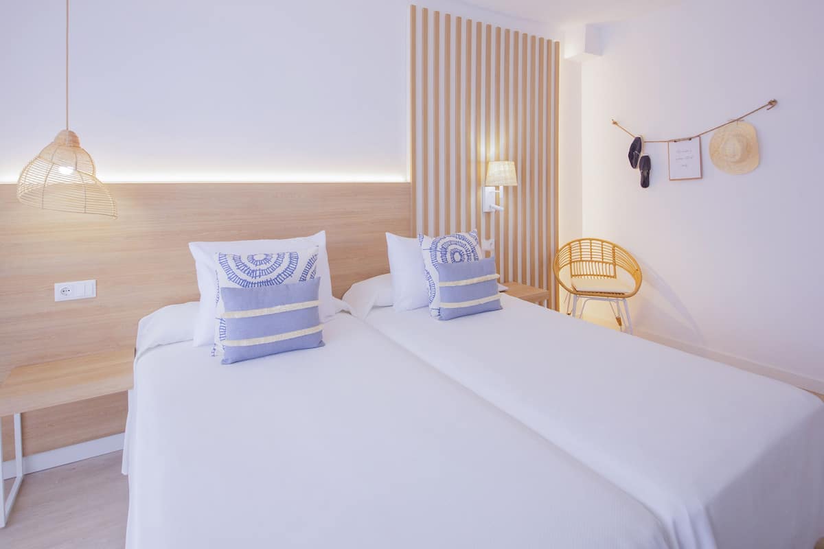 Habitación de hotel con dos camas, cabecero de madera, lámpara colgante, lámpara de pared, una silla de madera pequeña y un perchero de pared con un sombrero y un par de zapatos.
