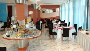 A spacious dining area with multiple round tables, each set with white tablecloths and black chairs. There are various food items and beverages displayed on a central island.