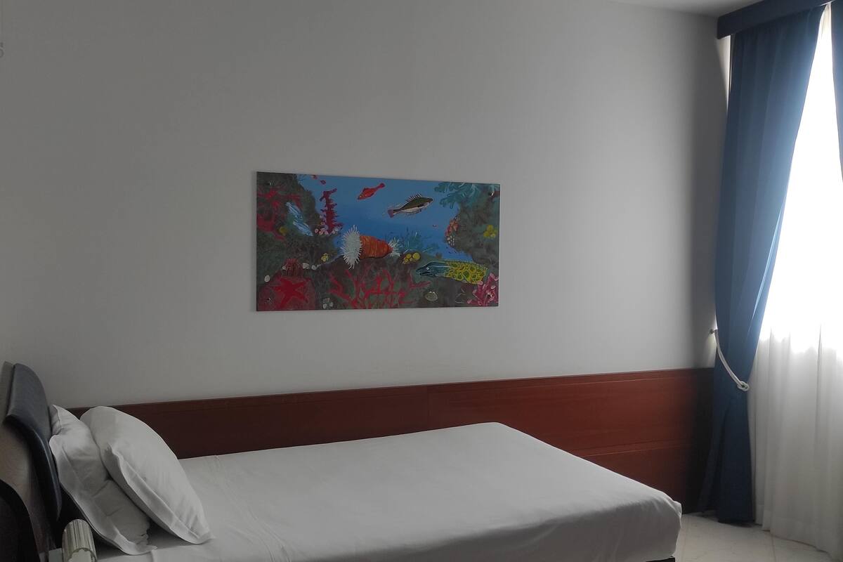 A hotel room with a bed, a wall-mounted painting, a window with curtains, and a wooden headboard.