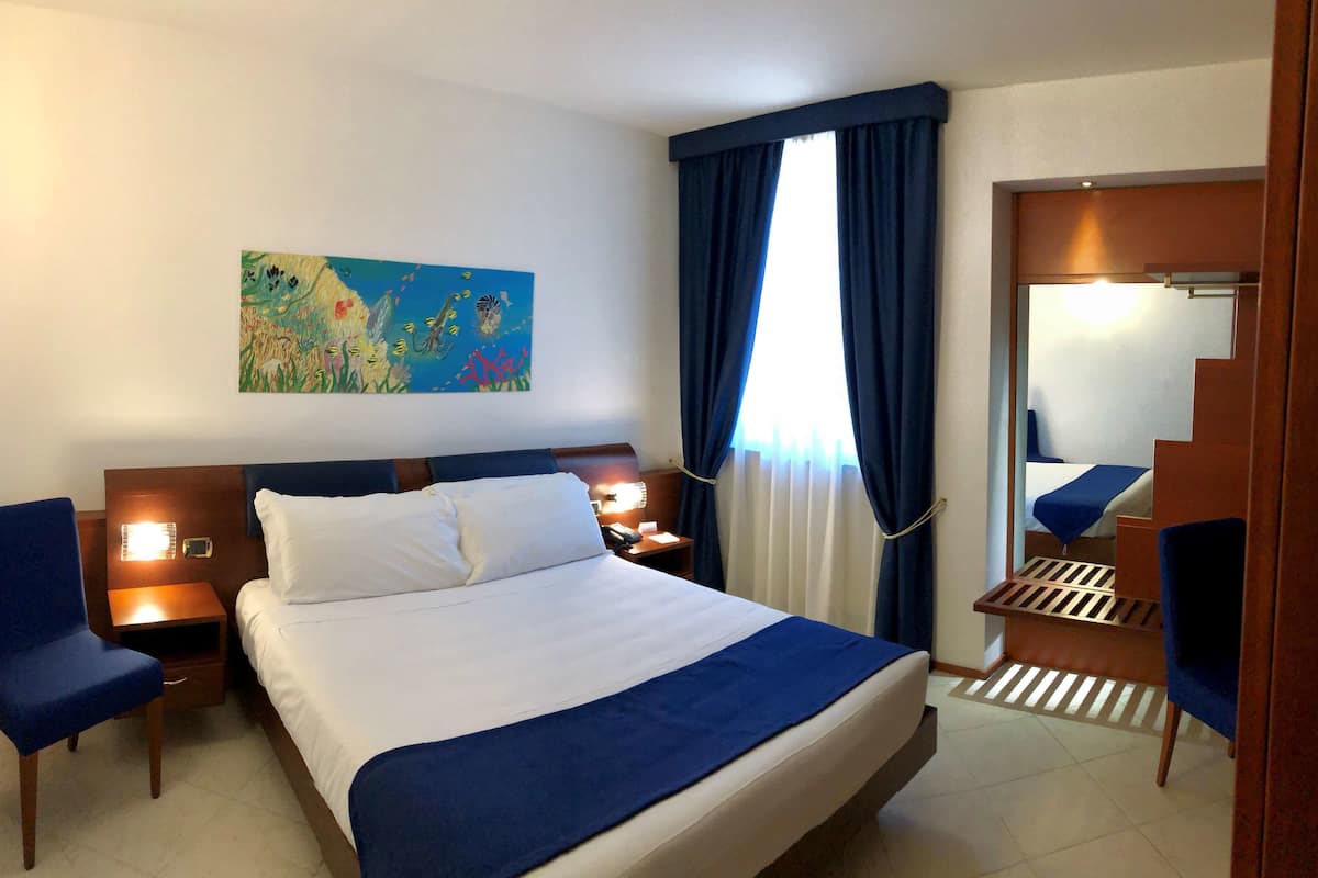 A hotel room with a bed, two bedside tables, a nightstand, a chair, a painting, a window with curtains, and a mirror.