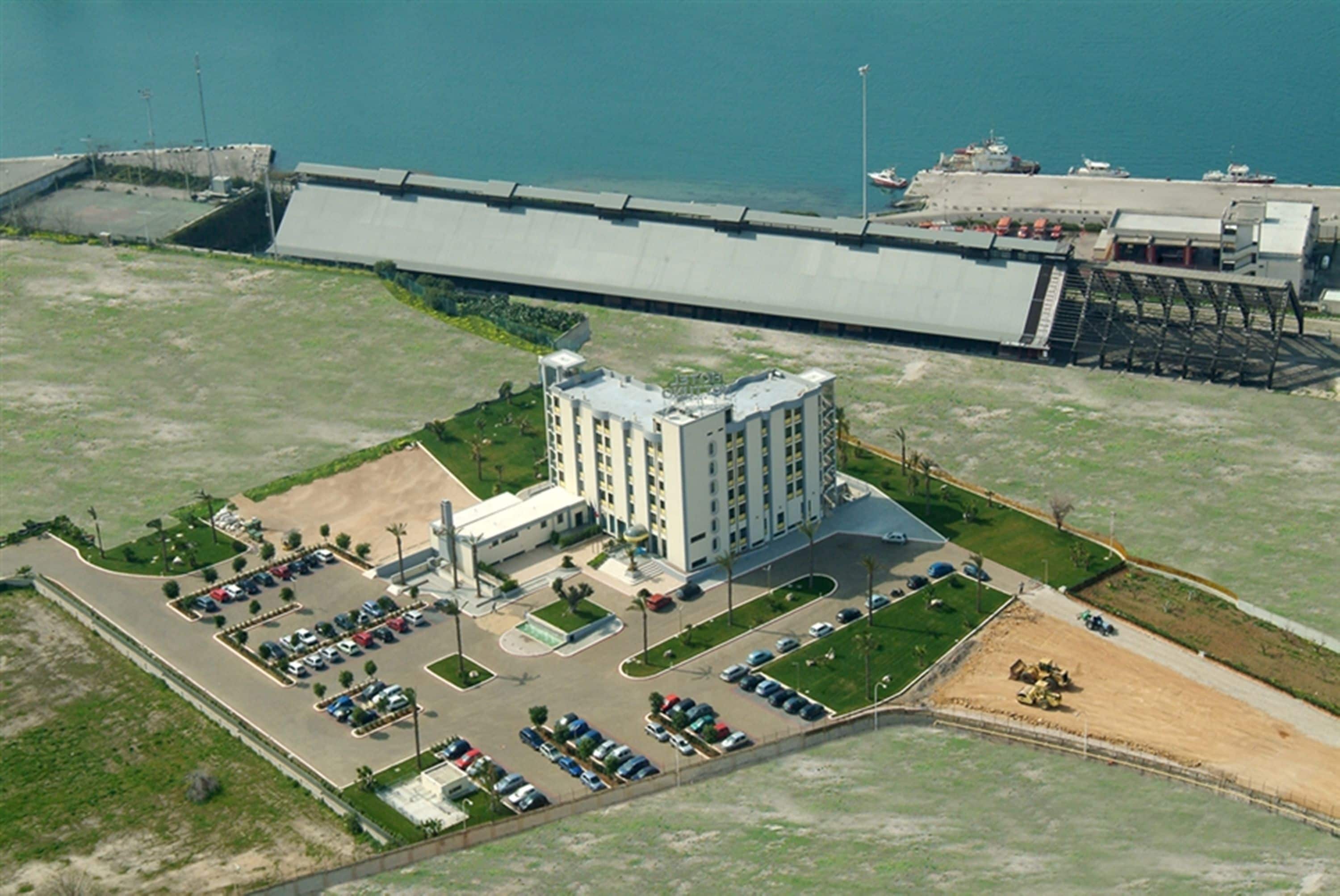 Vista aérea de un hotel frente al mar con estacionamiento, piscina y un edificio grande de techo plano.