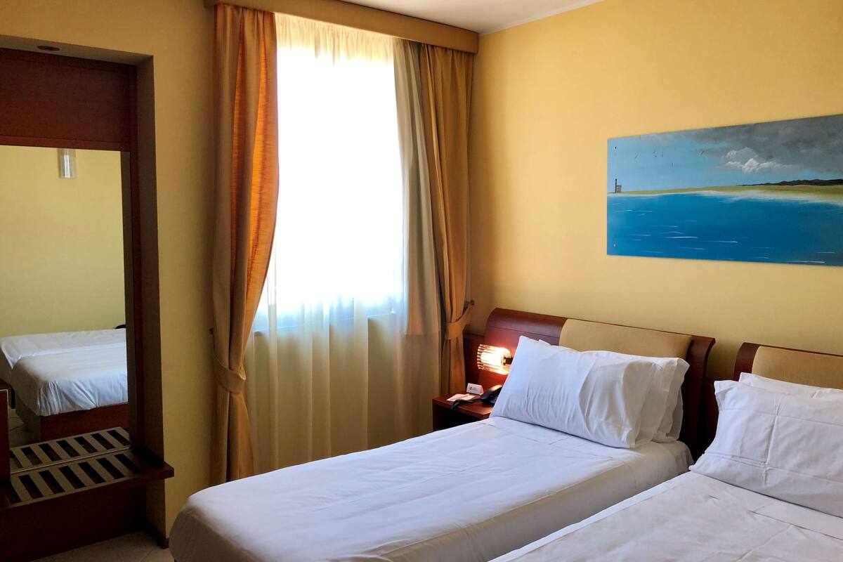 A hotel room with two beds, a painting of a coastal scene, and a window with curtains.