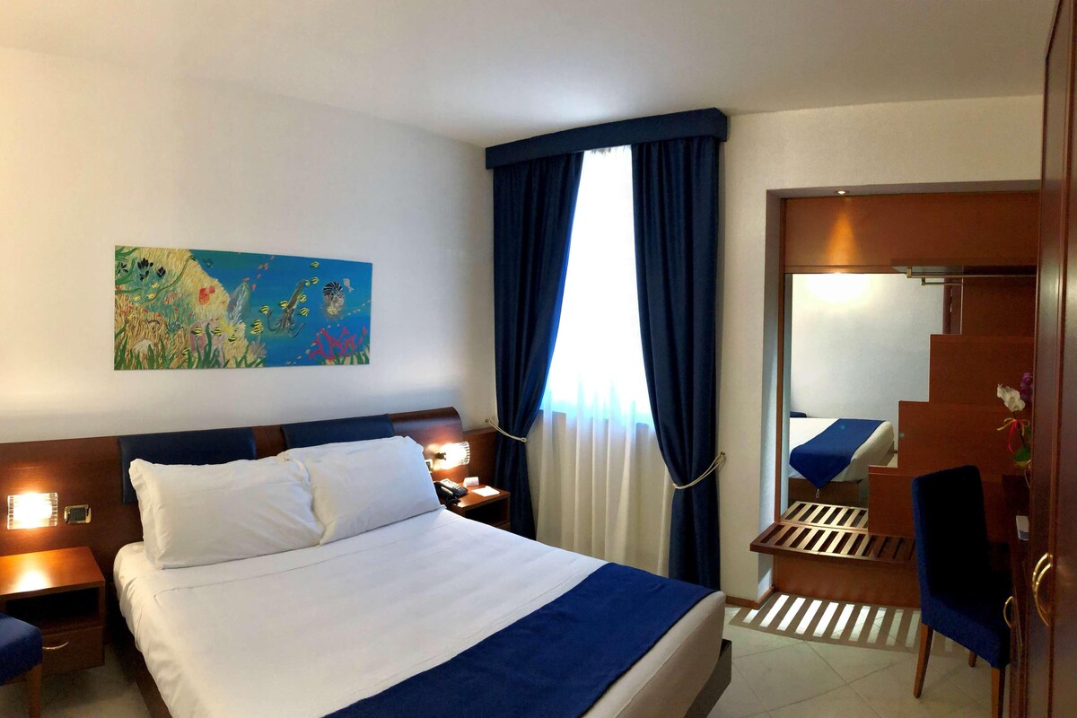 Una habitación de hotel con cama, mesitas de noche, escritorio, silla y un cuadro en la pared.