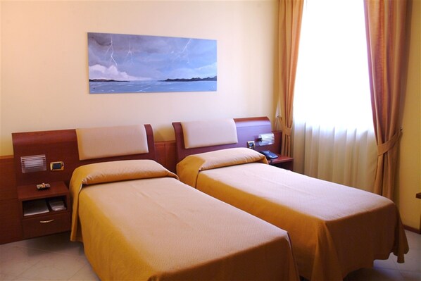 Habitación de hotel con dos camas individuales, una mesita de noche con lámpara, un cuadro en la pared y una ventana con cortinas.