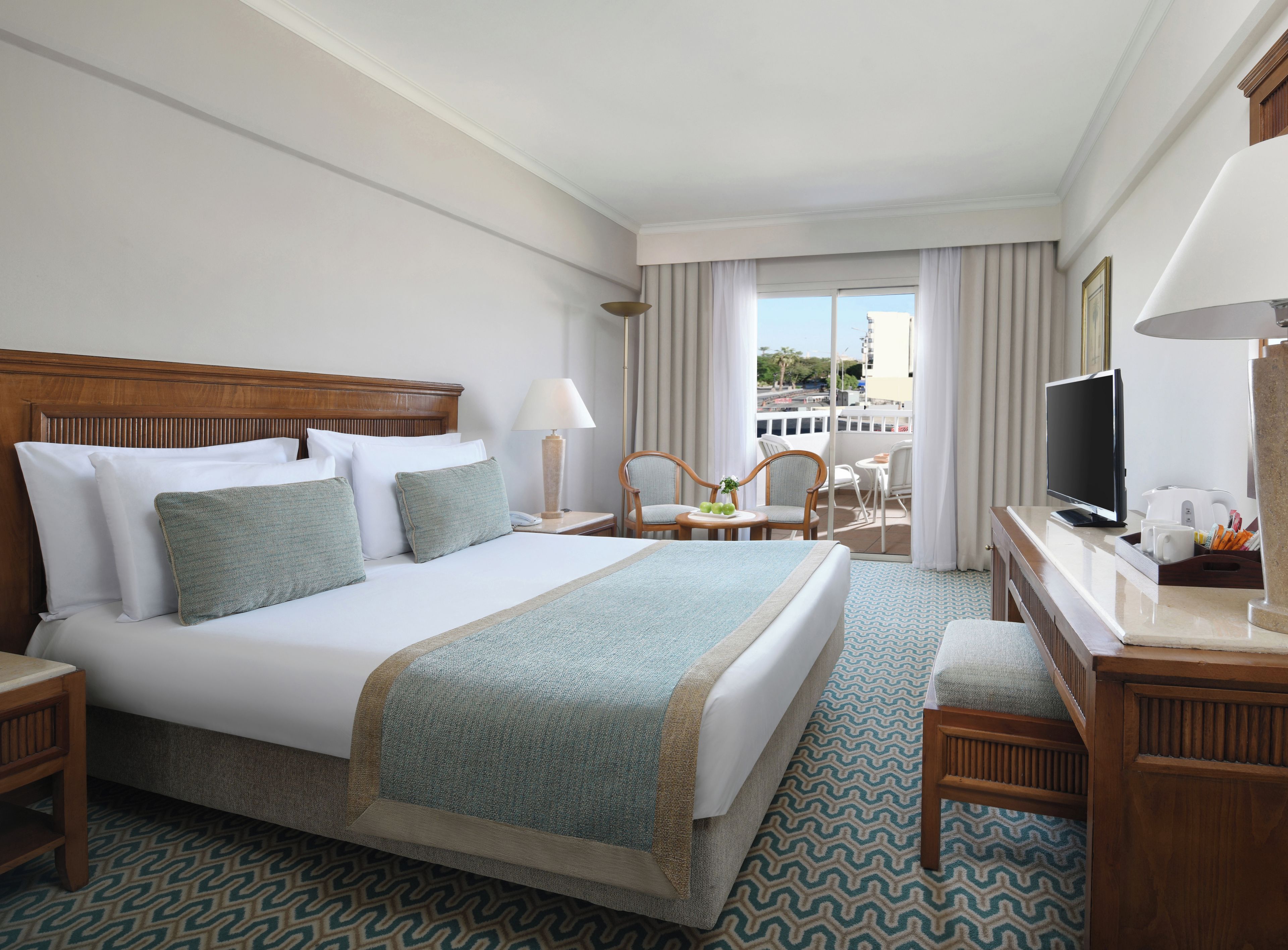 A hotel room with a large bed, a desk with a television, a chair, and a view of the city through the window.