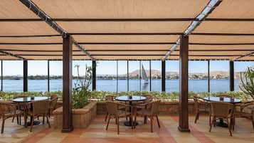 An outdoor patio with round tables and wicker chairs, overlooking a body of water with distant hills.