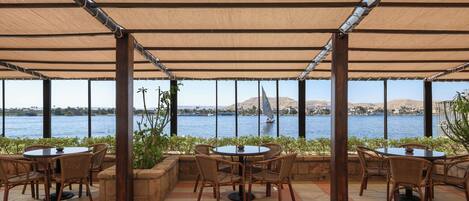 An outdoor patio with round tables and wicker chairs, overlooking a body of water with distant hills.