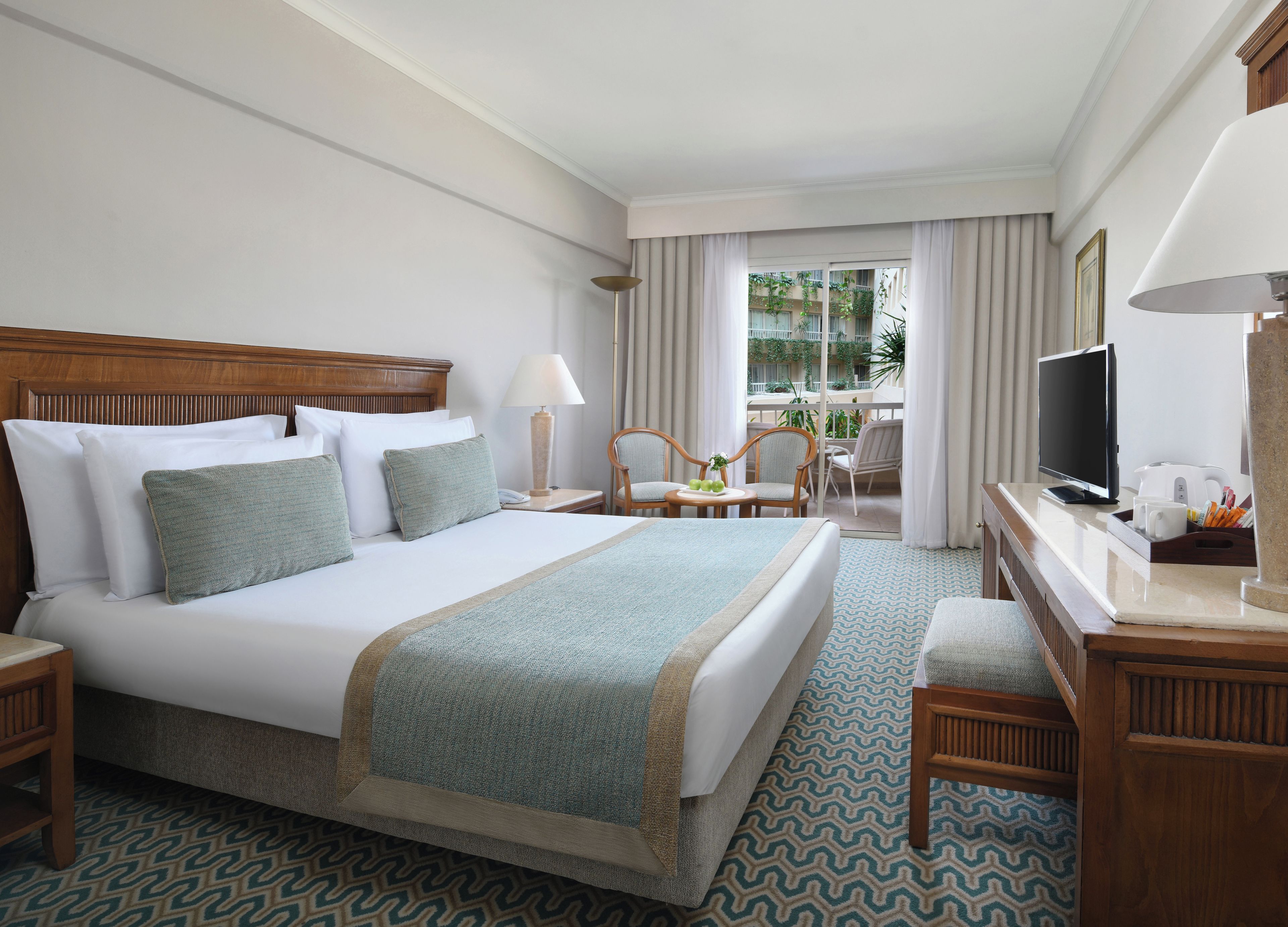A hotel room with a large bed, a desk with a television, a chair, and a view of a balcony with plants.