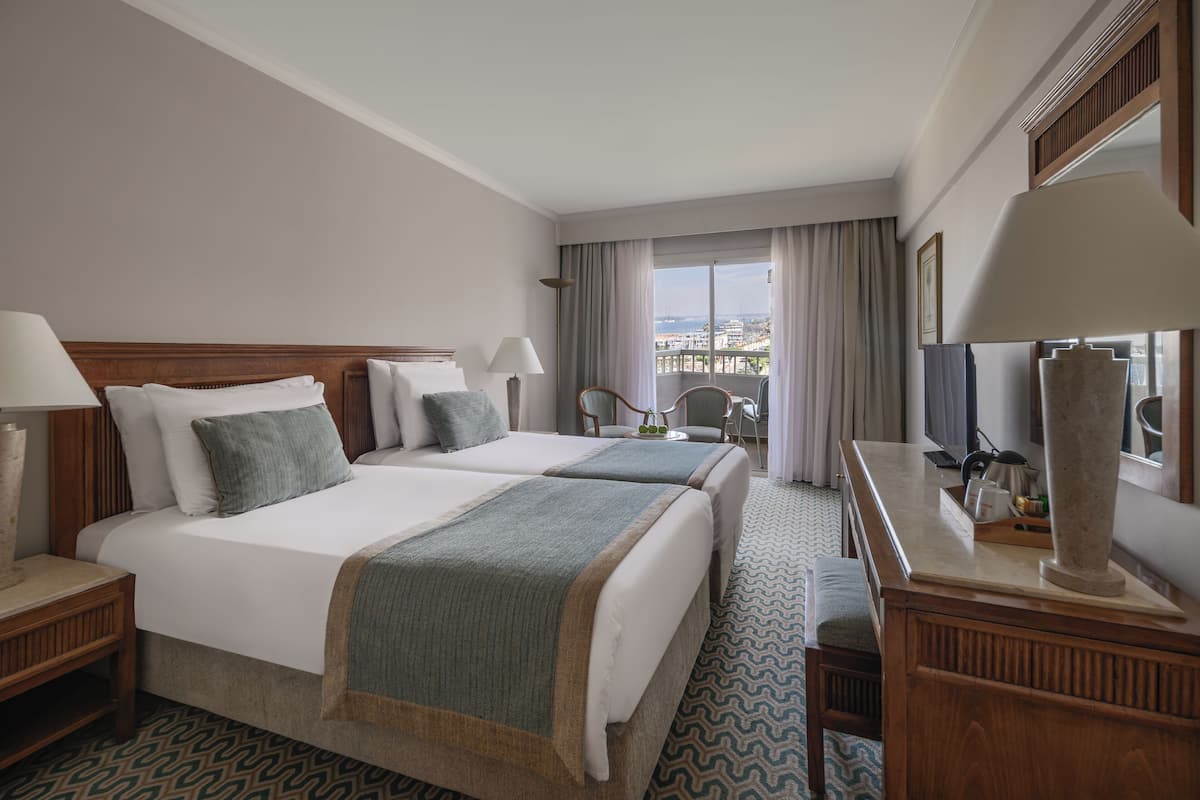 A hotel room with two beds, a desk with a lamp, a chair, a TV, and a view of the city.