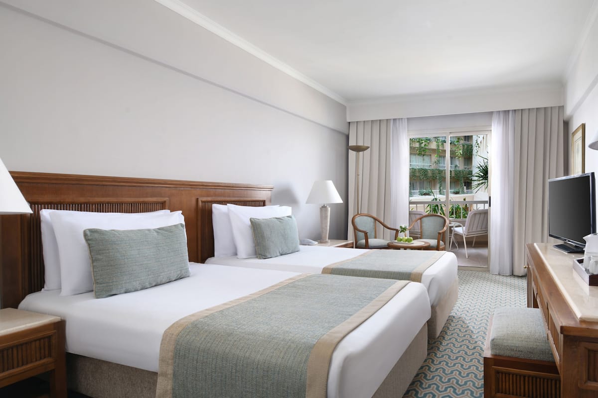 Two single beds with white linens and throw blankets, a wooden nightstand, and a round table with a vase of green apples.
