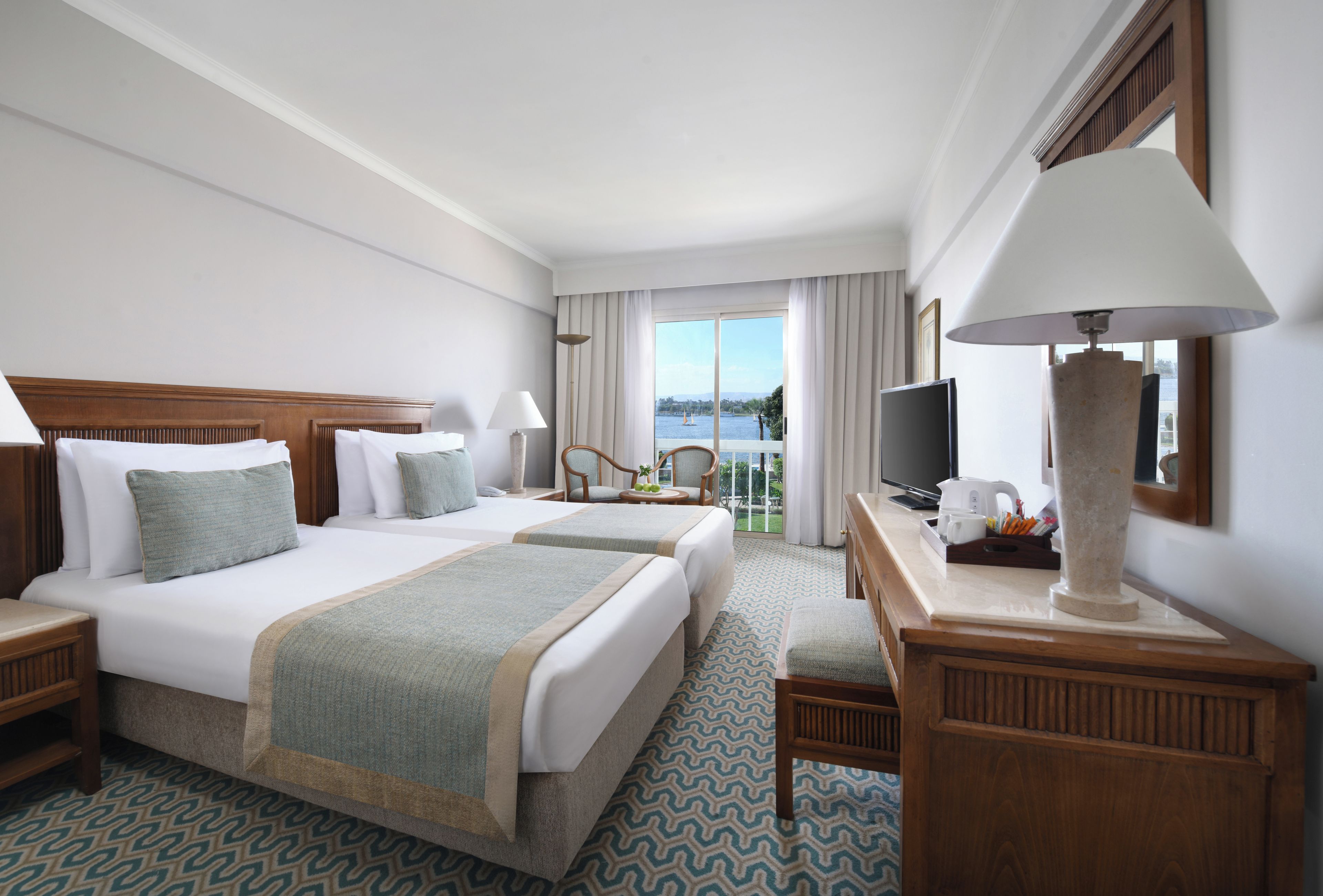 A hotel room with two beds, a television, a desk with a lamp, and a view of the sea.