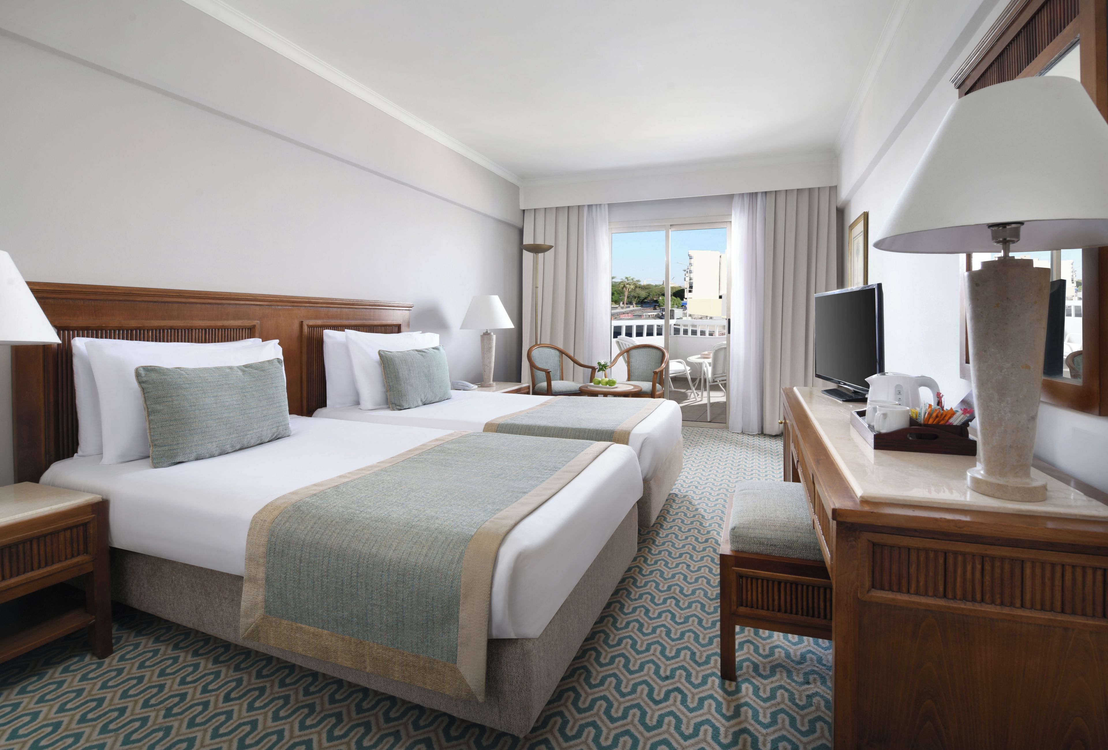 A hotel room with two beds, a desk, a television, and a balcony with a view.
