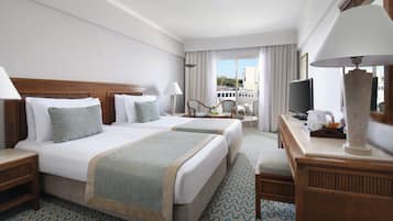 A hotel room with two beds, a desk, a television, and a balcony with a view.