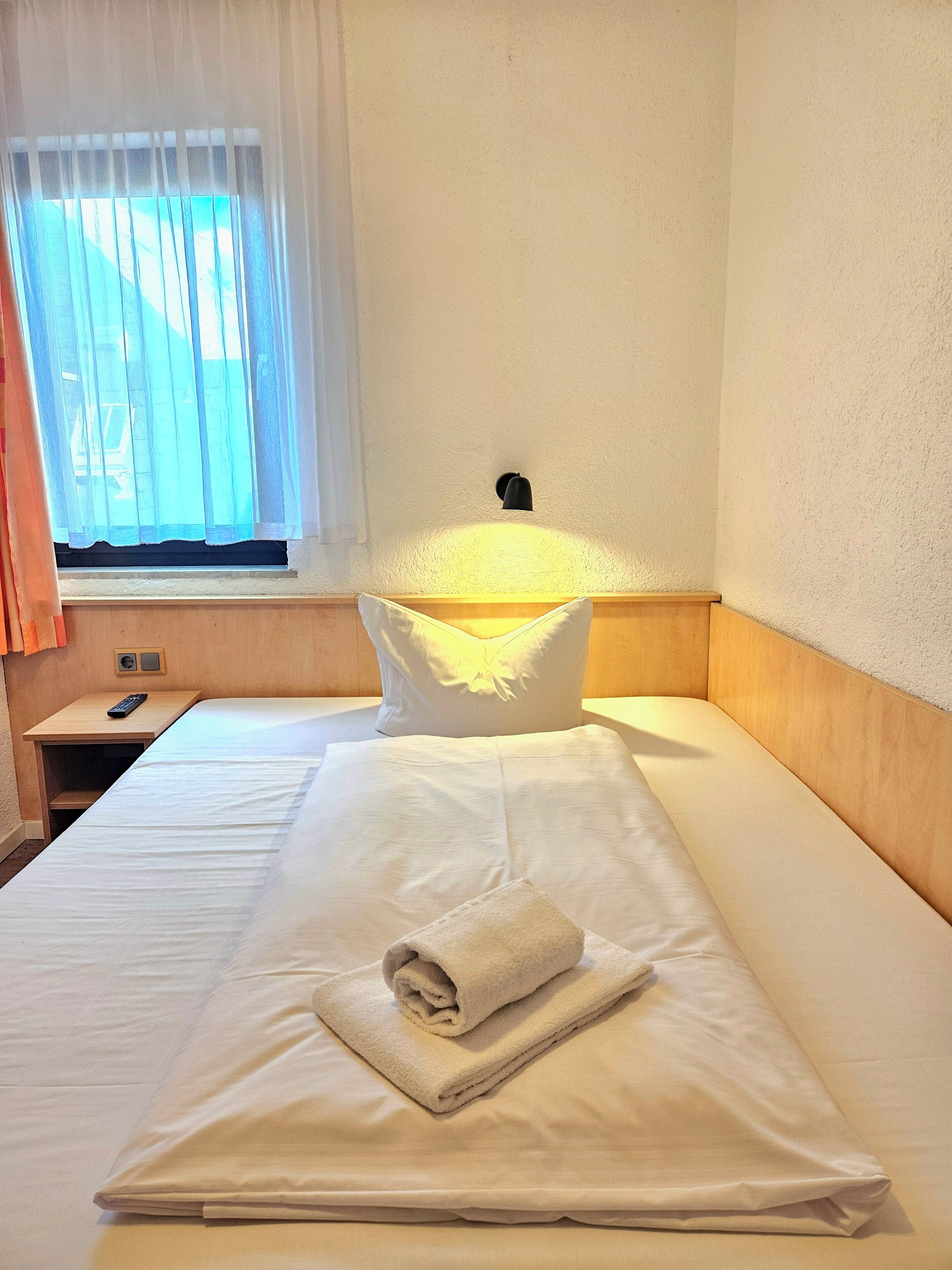 Una cama bien hecha con una toalla doblada, una mesita de noche con un control remoto y una ventana con cortinas traslúcidas.