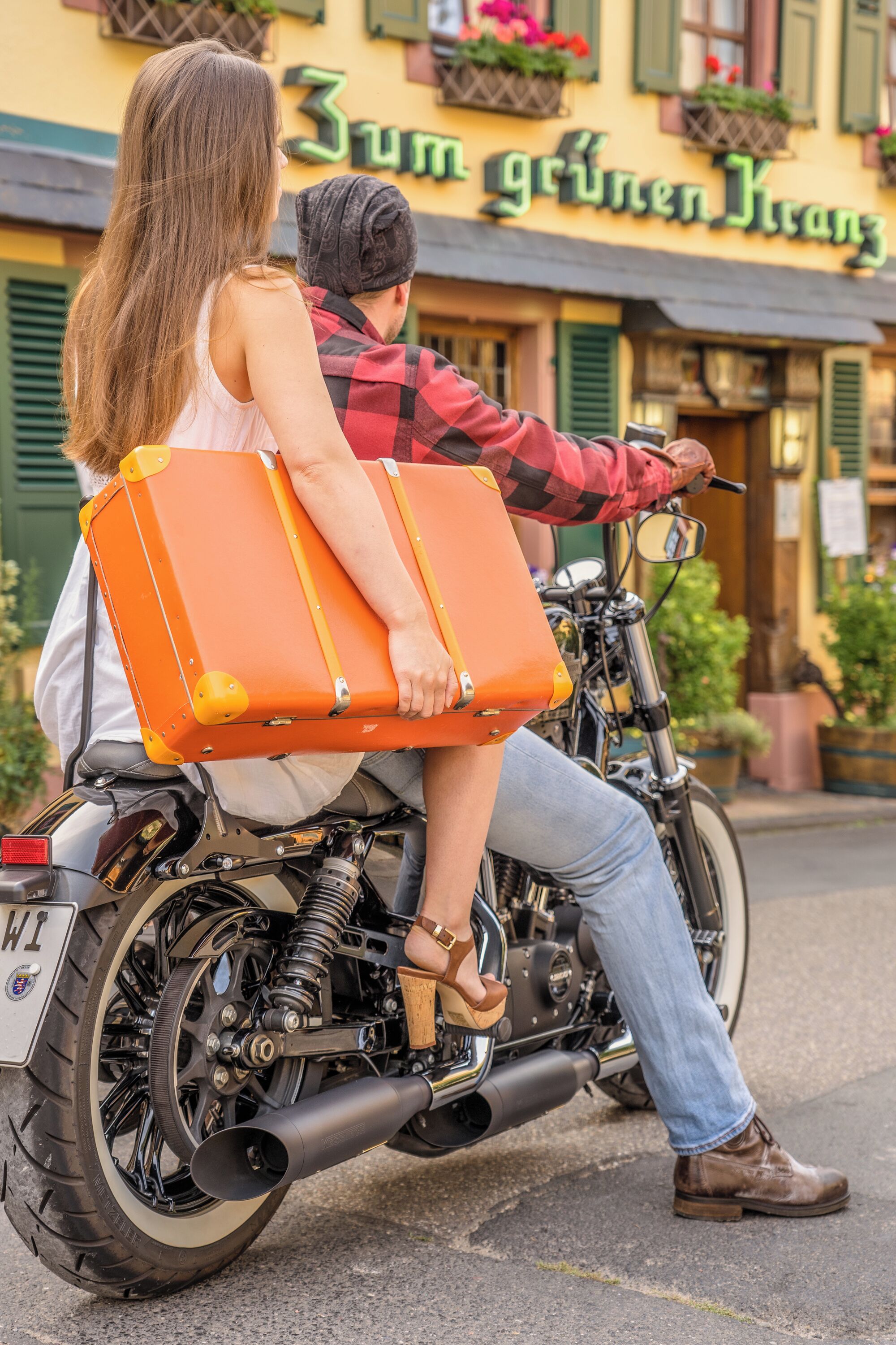 A couple on a motorcycle with a large orange suitcase, parked in front of a building with a sign reading "Zum grünen Kranz".
