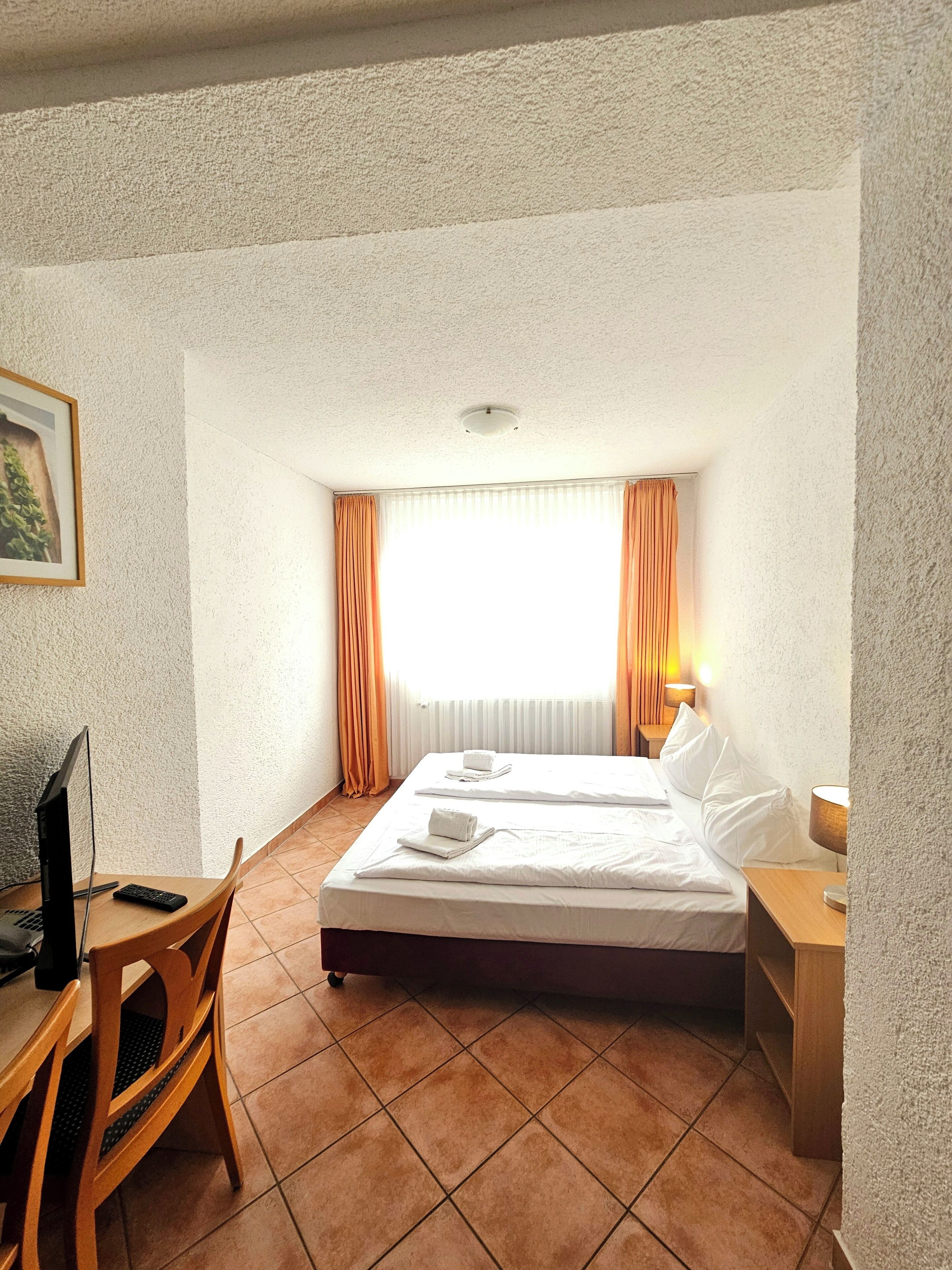 A hotel room with a bed, a desk with a computer, a chair, a window with curtains, and a lamp.