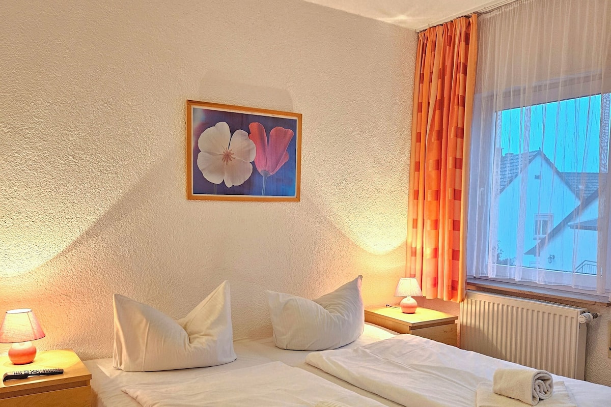 Een hotelkamer met twee bedden, een nachtkastje met een lamp, een schilderij aan de muur en een raam met gordijnen.