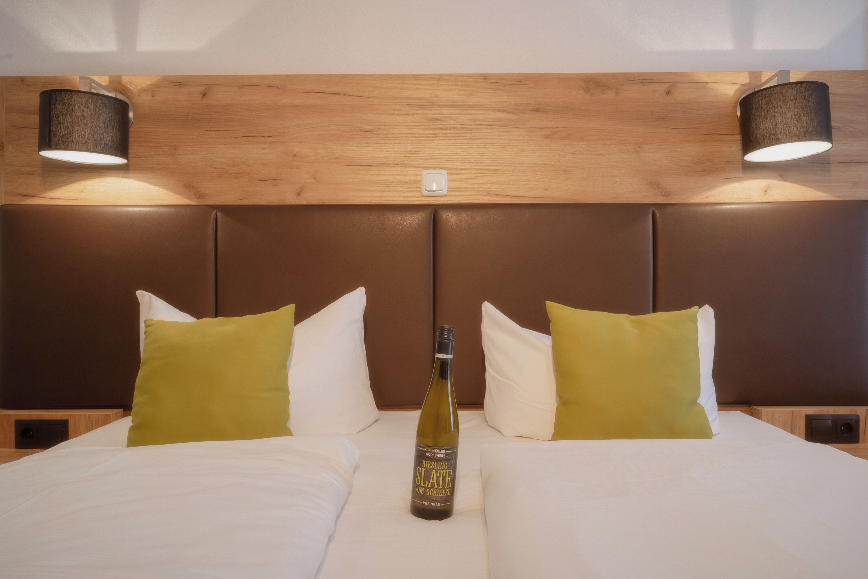 A hotel room with a wooden headboard, two beds with white and green pillows, and a bottle of wine on the bed.
