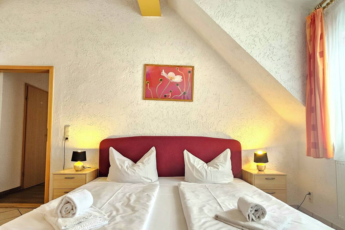 Een hotelkamer met een rode hoofdbord, twee bedden met witte lakens, een schilderij aan de muur en een deur naar een andere kamer.