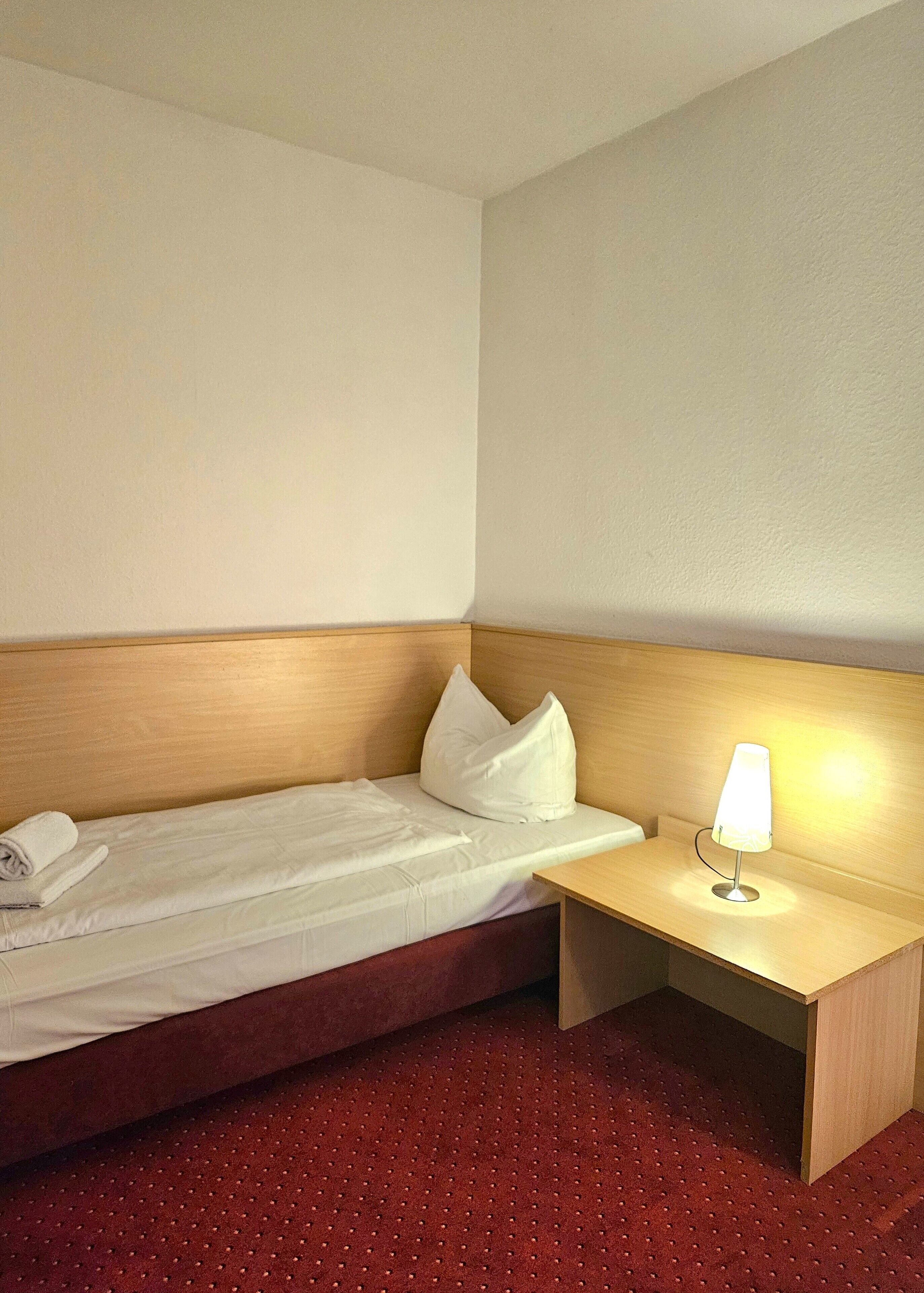Una habitación de hotel con una cama individual, cabecera de madera, mesita de noche con lámpara y suelo cubierto de alfombra roja.