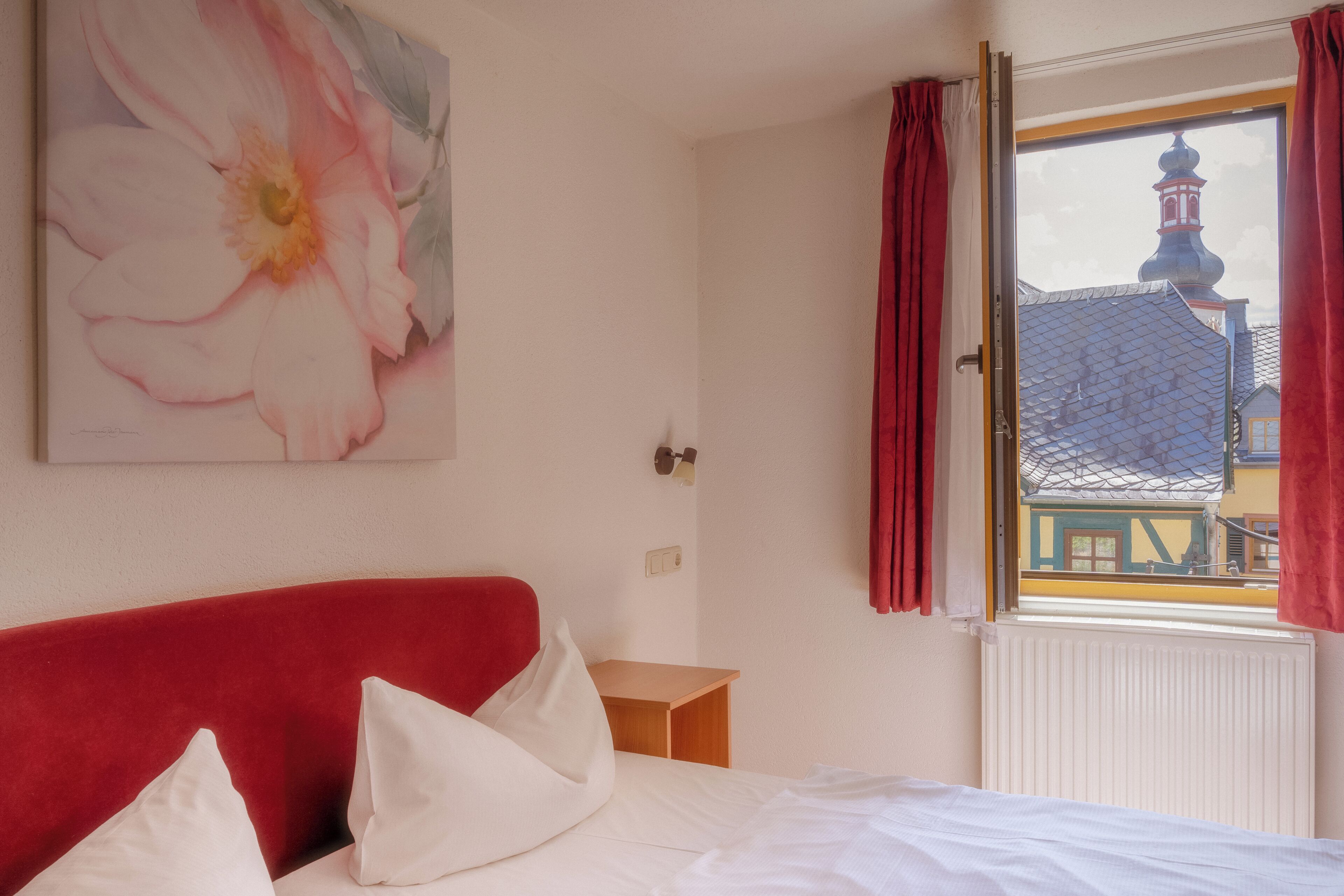 Et hotelværelse med en seng, et trænatbord, en rød sengegavl, et vindue med røde gardiner og et maleri af en blomst på væggen.