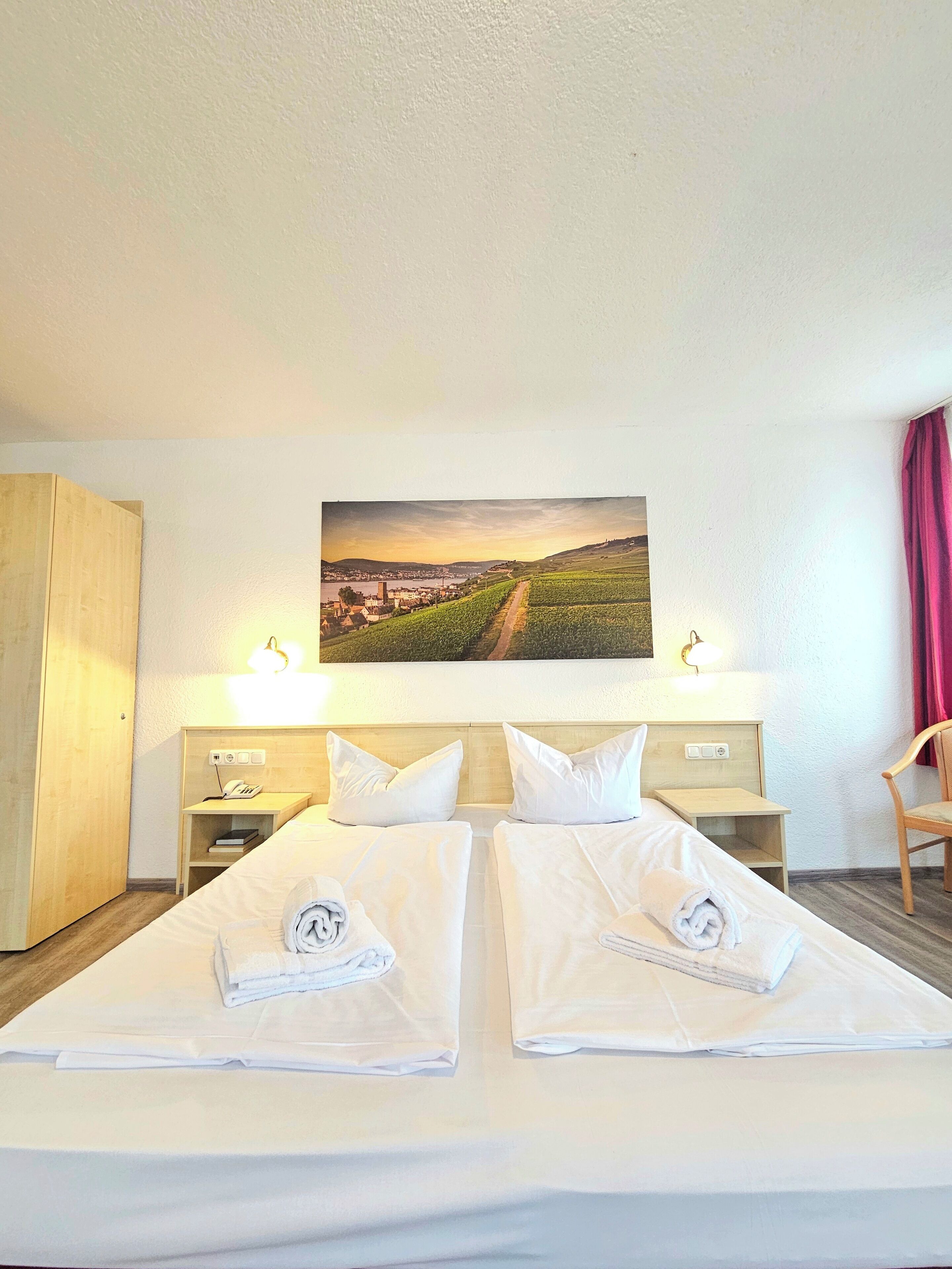Habitación de hotel con dos camas, un cuadro de un viñedo, lámparas de noche y un armario de madera.