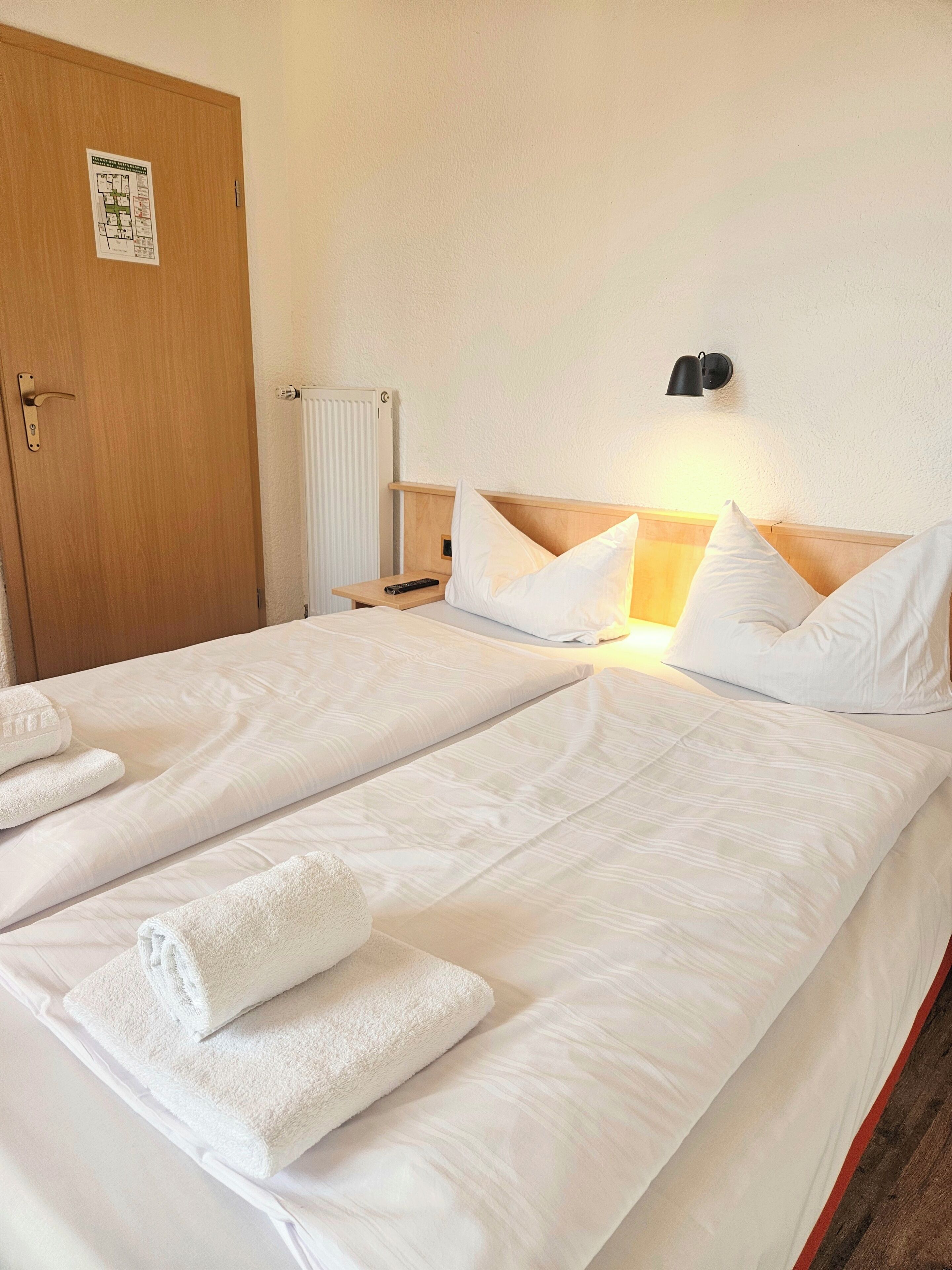 A hotel room with two beds, white bedding, a wooden headboard, a nightstand, a wall-mounted lamp, and a radiator.