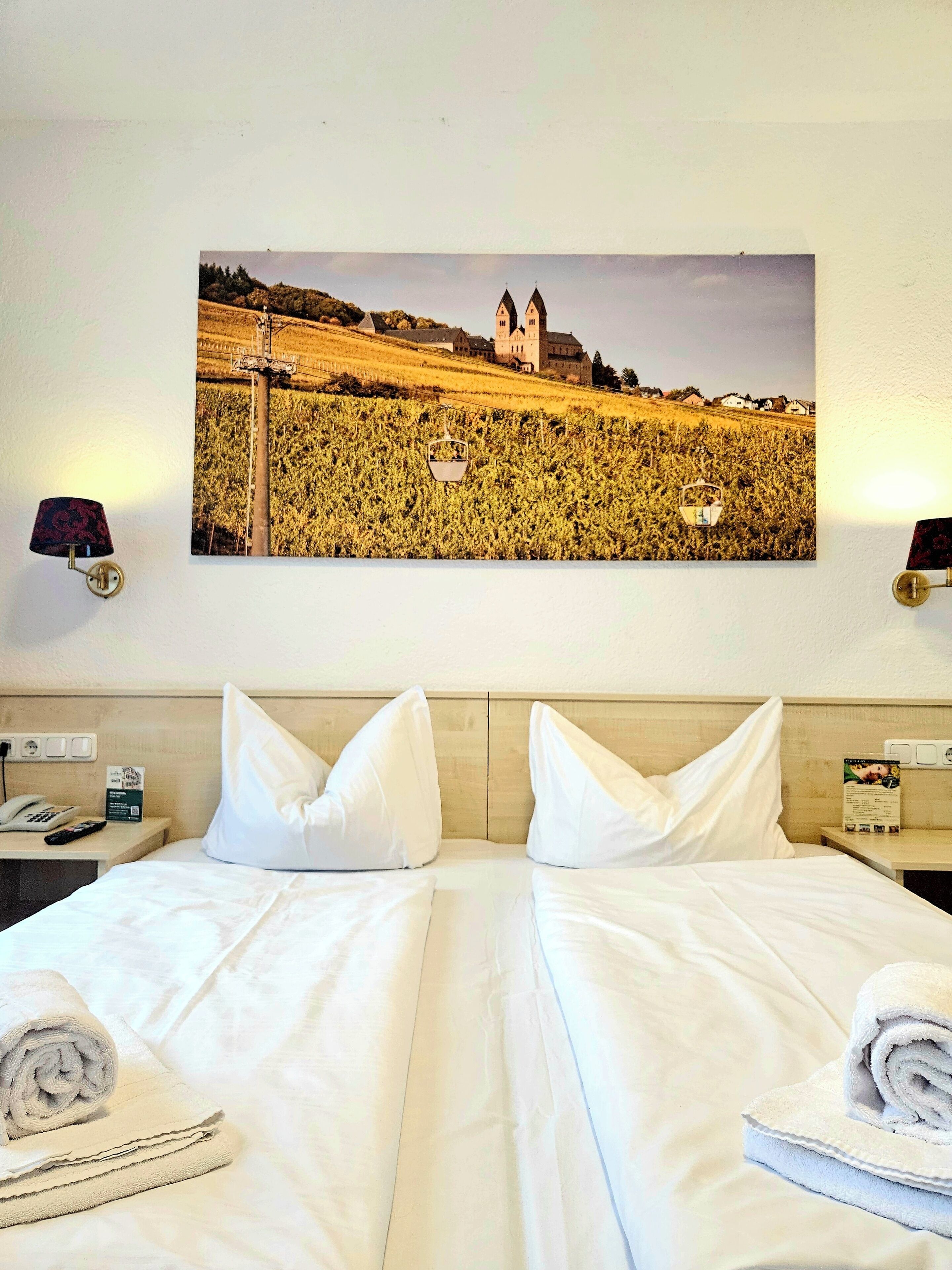 A hotel room with a large bed, two pillows, a nightstand with a lamp, a remote control, a folded towel, and a framed picture of a castle on the wall.