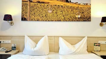 A hotel room with a large bed, two pillows, a nightstand with a lamp, a remote control, a folded towel, and a framed picture of a castle on the wall.