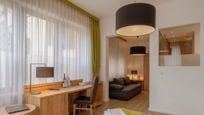 Habitación de hotel con escritorio de madera, una silla, cama con manta verde, ventana grande con cortinas translúcidas y una lámpara colgante moderna.