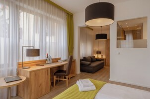 Habitación de hotel con escritorio de madera, una silla, cama con manta verde, ventana grande con cortinas translúcidas y una lámpara colgante moderna.