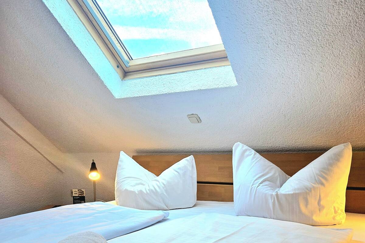 Een opgemaakt bed met witte lakens en kussens, een opgerolde handdoek en een kleine tafellamp.