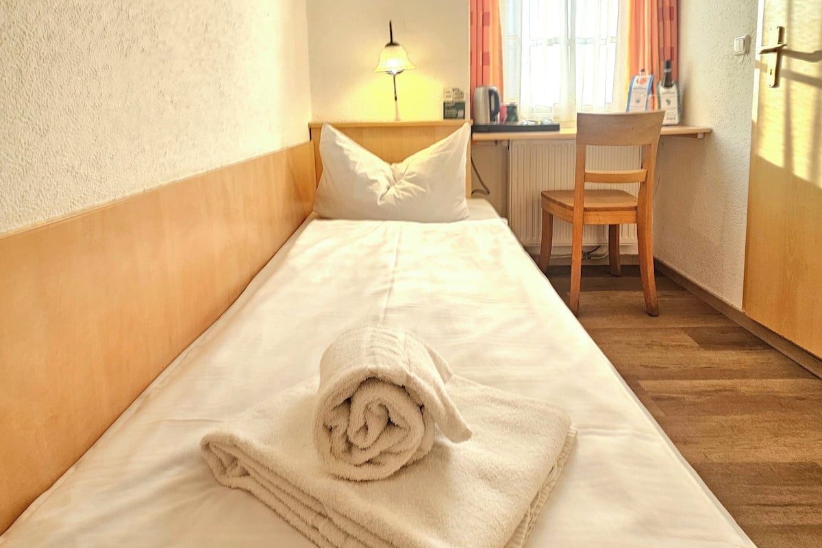 Een net opgemaakt bed met een opgerolde handdoek, een bedlamp, een stoel en een raam met gordijnen.