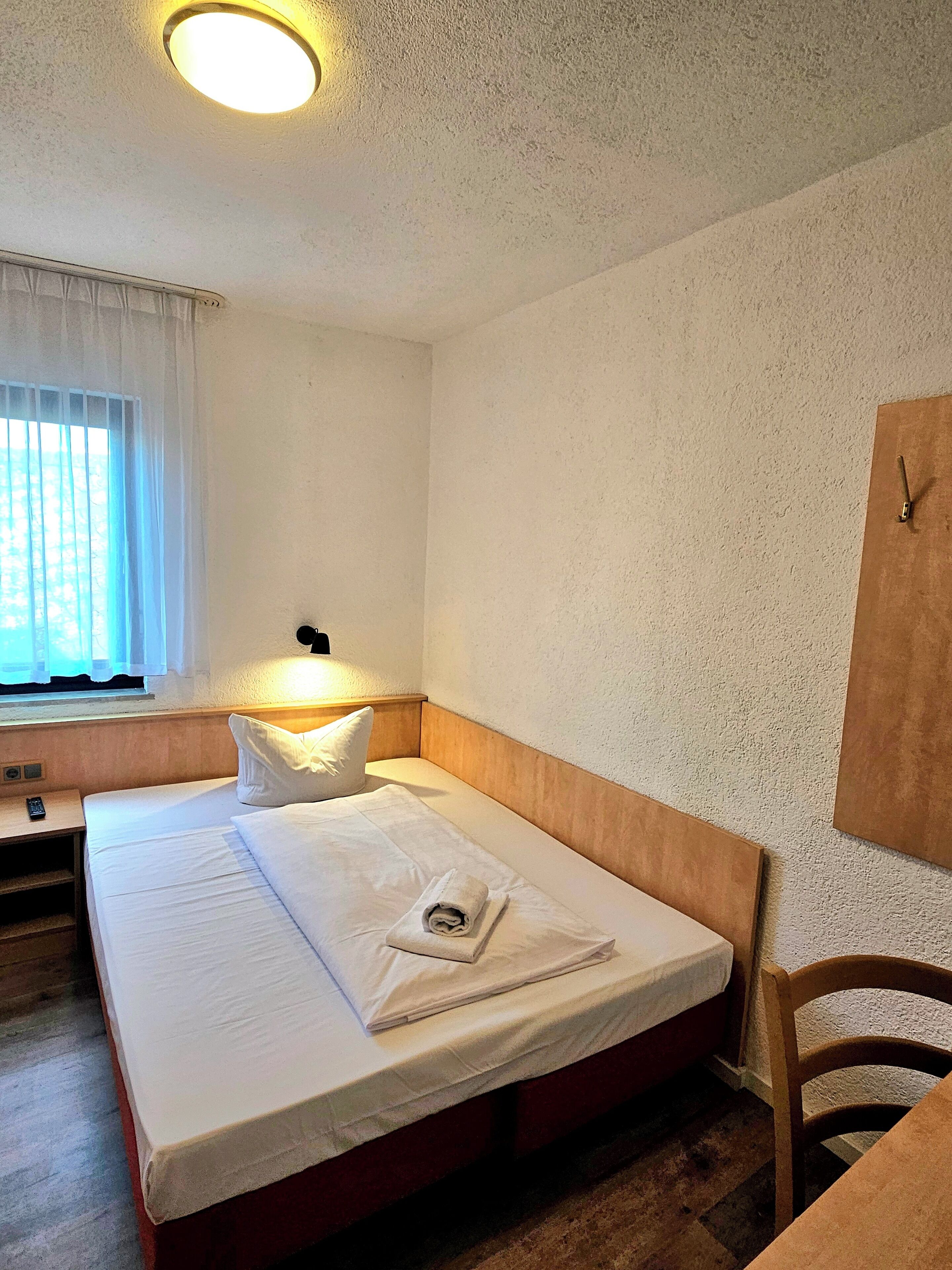 Une chambre d’hôtel avec un lit simple, une table de chevet avec une télécommande, une fenêtre avec des rideaux et une horloge fixée au mur.