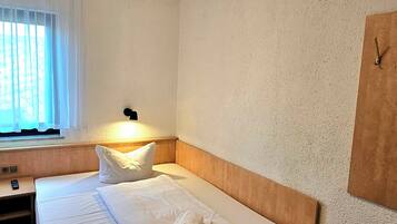 Une chambre d’hôtel avec un lit simple, une table de chevet avec une télécommande, une fenêtre avec des rideaux et une horloge fixée au mur.