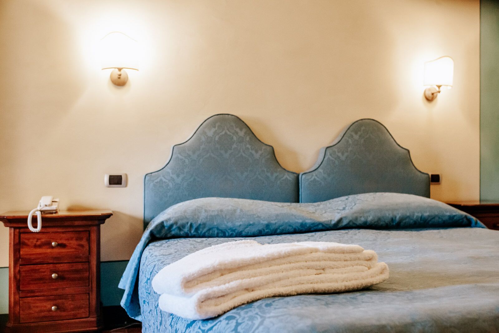 A neatly made bed with a folded white towel, a wooden nightstand with a telephone, and two wall-mounted light fixtures.
