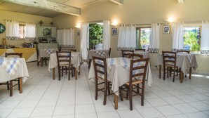 Un comedor con varias mesas y sillas, una cocina con armarios y un refrigerador, y ventanas con cortinas.