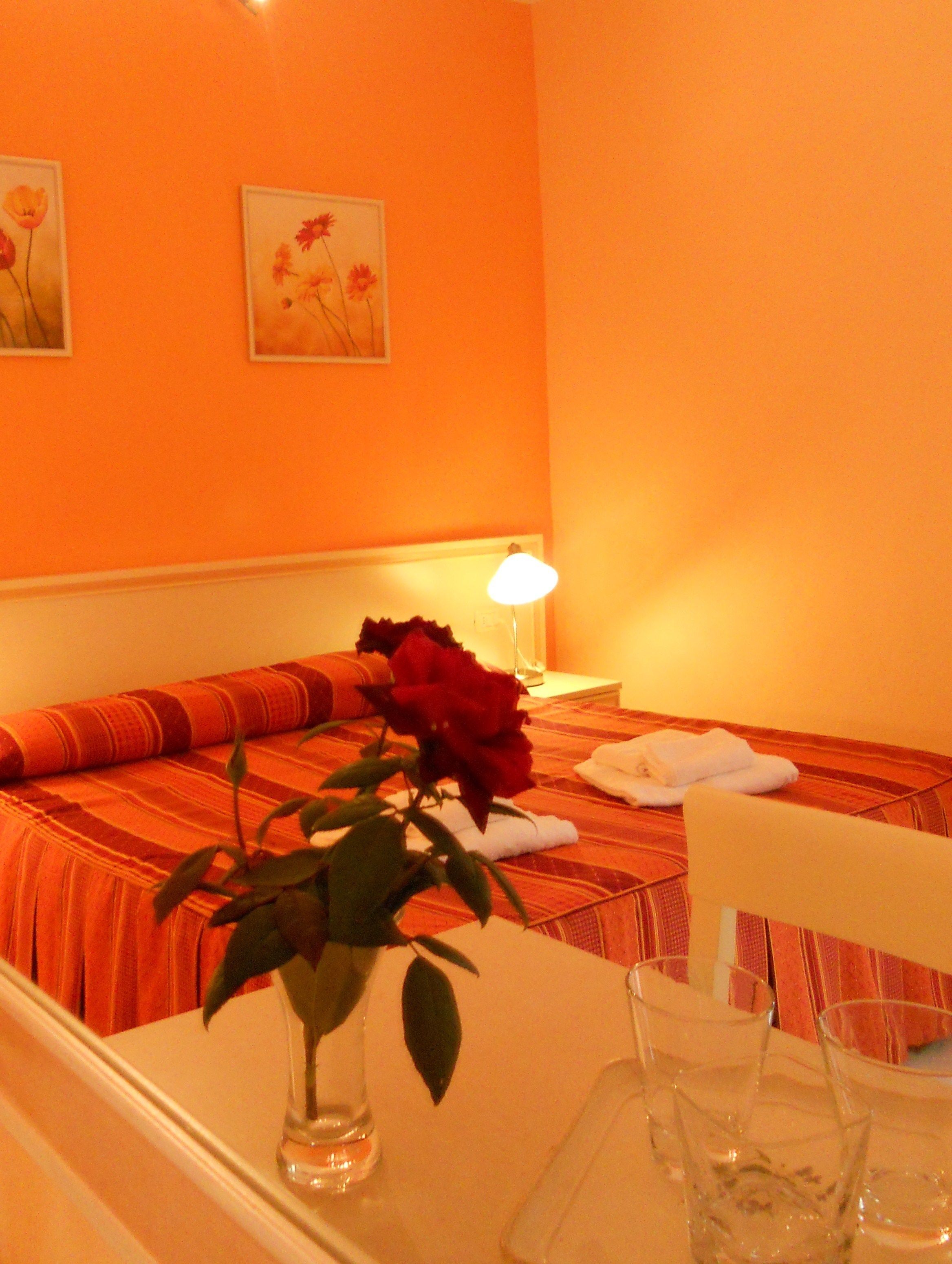 Quarto de hotel com uma cama, uma mesa de cabeceira com um vaso de flores e uma luminária.