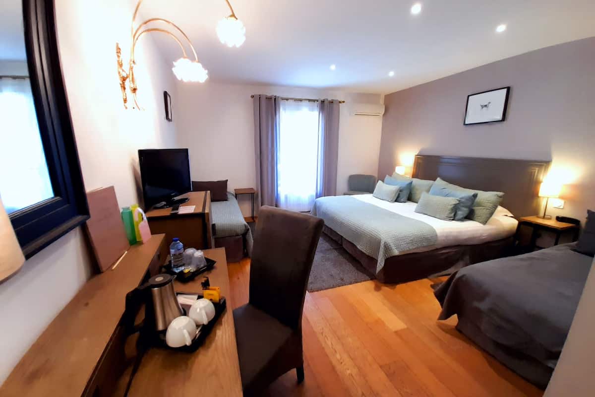 A hotel room with a bed, a desk with a TV, a chair, a coffee maker, and a bedside table with a lamp.