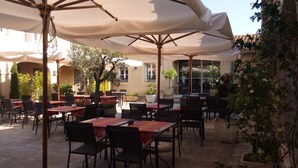An outdoor café with tables and chairs, covered by large umbrellas, and surrounded by potted plants and trees. - Hôtel Le Fabian des Baux (Les Baux-de-Provence)