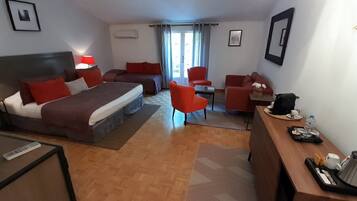 A hotel room with a bed, two red chairs, a desk with a coffee maker, and a view of the outdoors through a window.