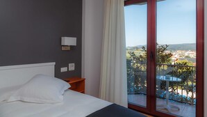 A hotel room with a bed, a nightstand, a wall-mounted lamp, and a balcony with a view of the city. - Hotel Lombiña (Puebla del Caraminal)