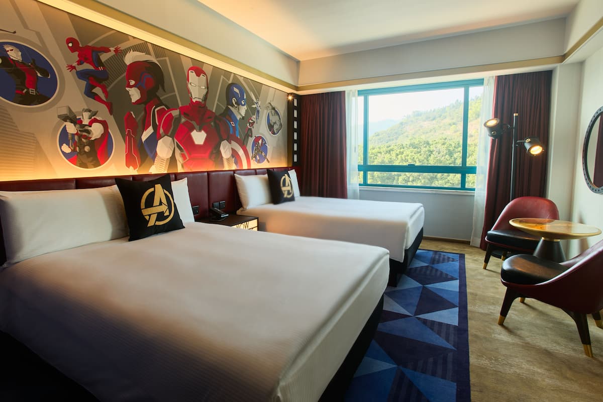 A hotel room with two beds, a large mural of superheroes, a view of a forest, and a red chair.