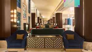 Un hall d’entrée spacieux avec un espace salon central comprenant un canapé vert et deux fauteuils bleus, une table basse à motifs et un panneau décoratif mural.