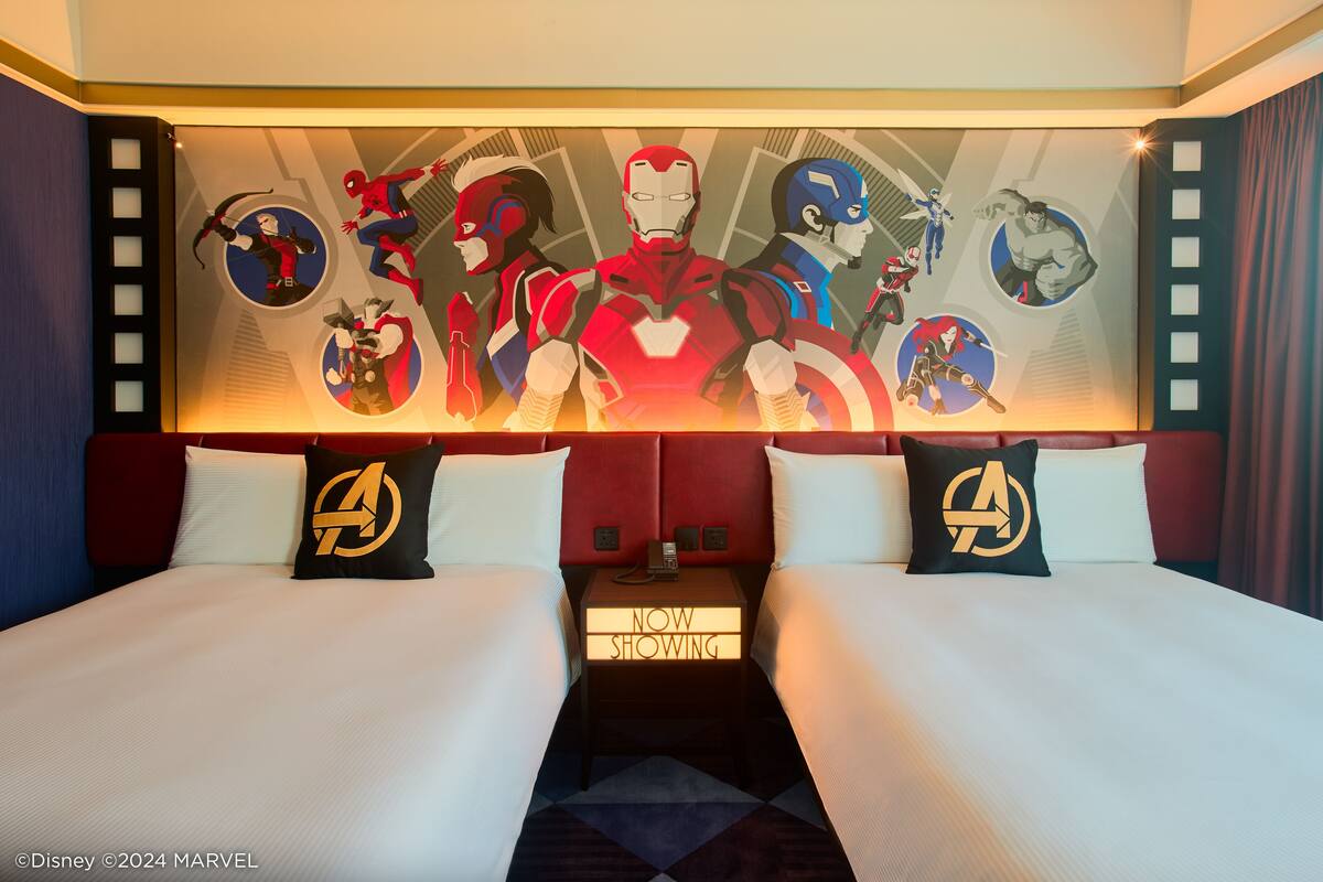 A hotel room with a large wall mural of Marvel superheroes, two beds with matching pillows, and a nightstand with a 'NOW SHOWING' sign.