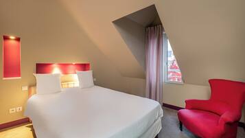 Ein Hotelzimmer mit einem großen Bett, einem roten Sessel, einer schrägen Decke und einem Fenster mit Vorhängen.